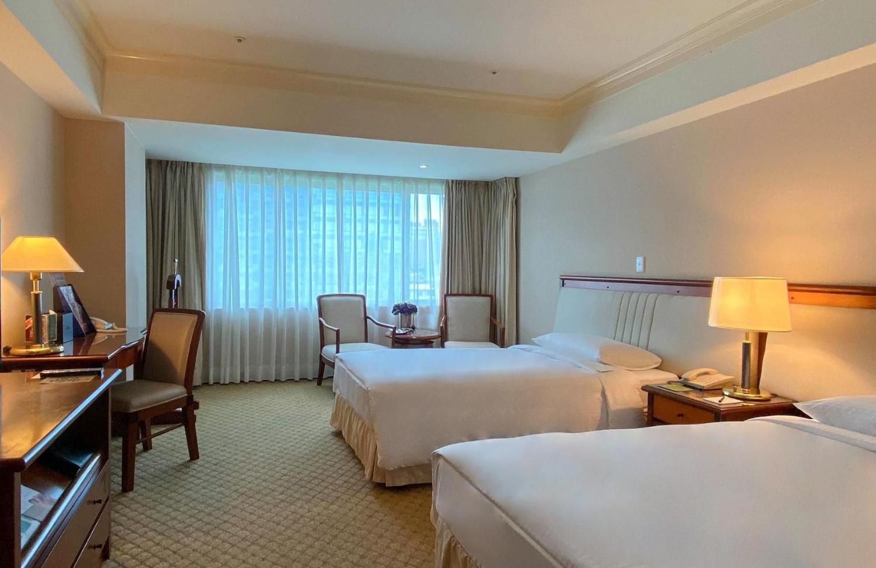 Evergreen Laurel Hotel - Taizhong Zewnętrze zdjęcie The photo shows a hotel room featuring two single beds positioned near each other. There's a light-colored carpet on the floor and a large window with sheer curtains that allows natural light to fill the space. In the room, there are two chairs place