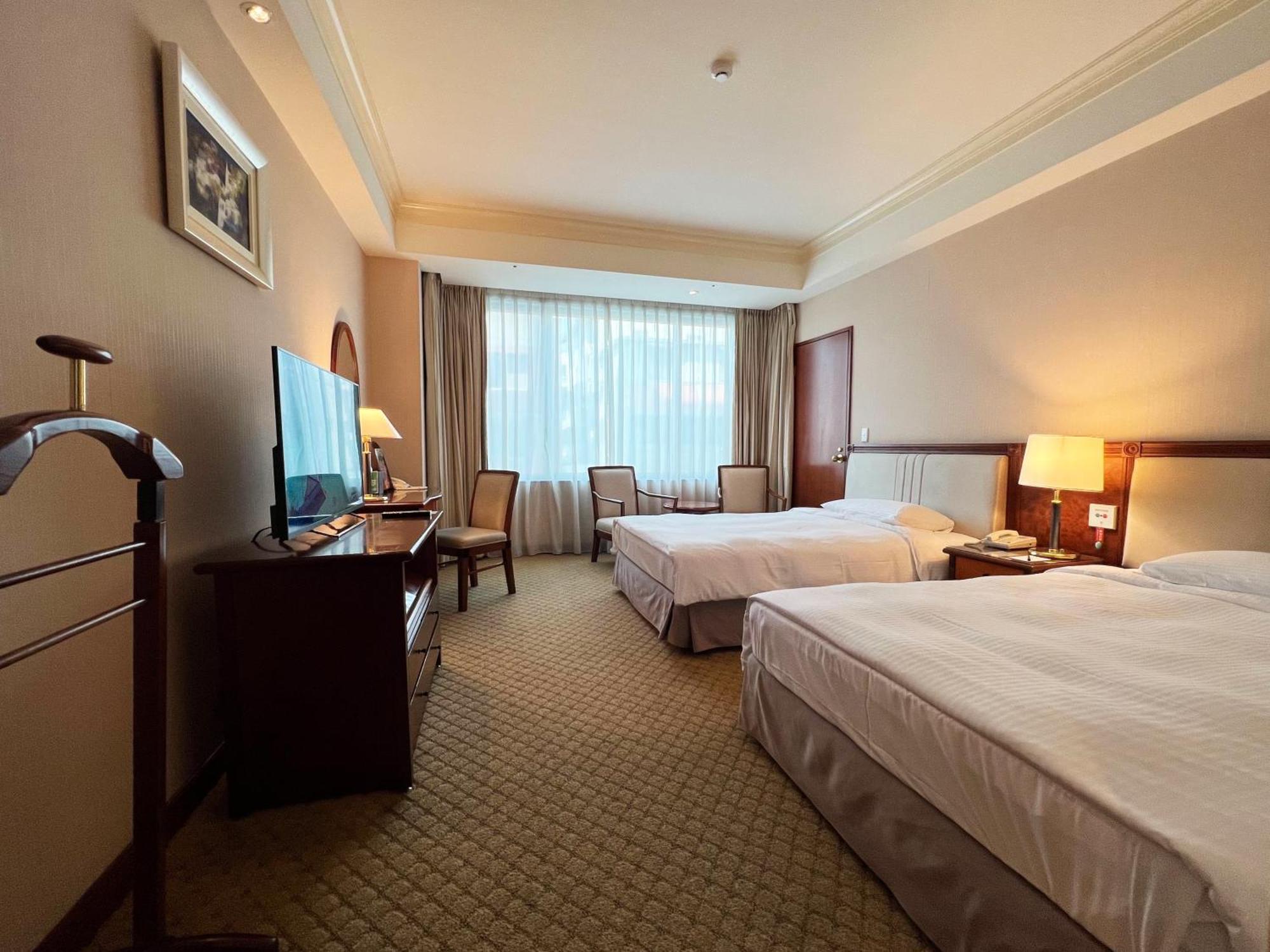 Evergreen Laurel Hotel - Taizhong Zewnętrze zdjęcie The photo shows a hotel room featuring two twin beds placed parallel to each other. The room has a warm color palette and includes a large window with sheer curtains allowing natural light to illuminate the space. There is a wooden desk and two chair