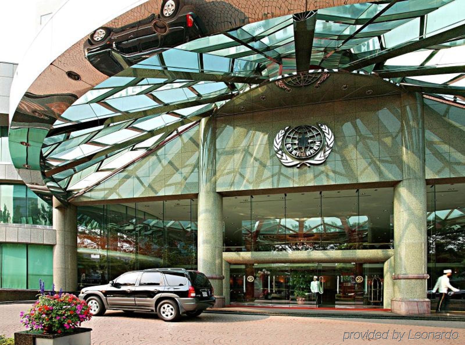 Evergreen Laurel Hotel - Taizhong Zewnętrze zdjęcie The photo shows the entrance of a modern building characterized by its glass and metal architecture. The structure features a large, domed roof that reflects light, with a distinctive emblem or logo displayed prominently at the center. In front of th