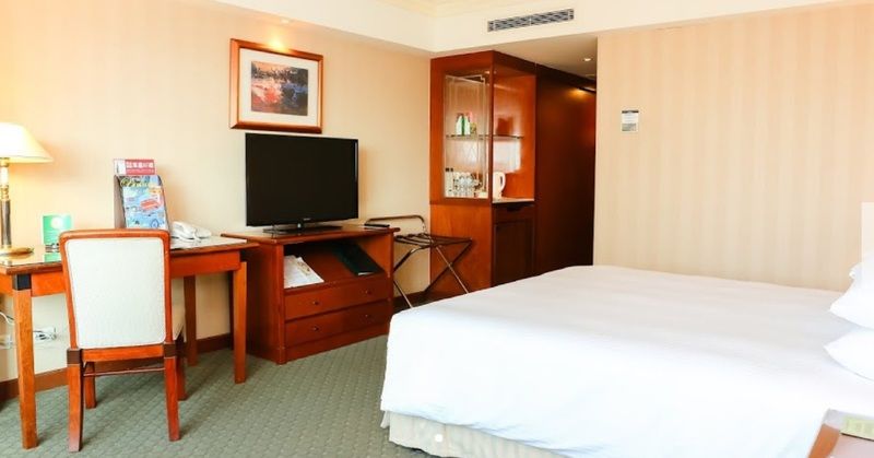 Evergreen Laurel Hotel - Taizhong Zewnętrze zdjęcie The photo shows a hotel room featuring a neatly made bed with a white blanket. On one side, there is a wooden desk with a chair, and a flat-screen television is mounted nearby. An accent cabinet is visible, displaying various items. The walls are pai