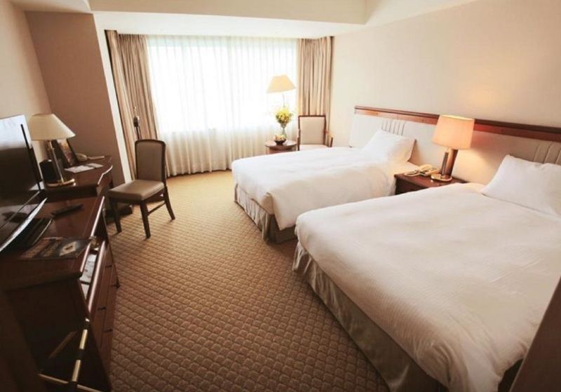 Evergreen Laurel Hotel - Taizhong Zewnętrze zdjęcie The photo shows a hotel room featuring two twin beds with white bedding. There's a chair positioned near one of the beds and a desk with a computer setup against one wall. Large windows allow natural light to enter the room, and light-colored curtain