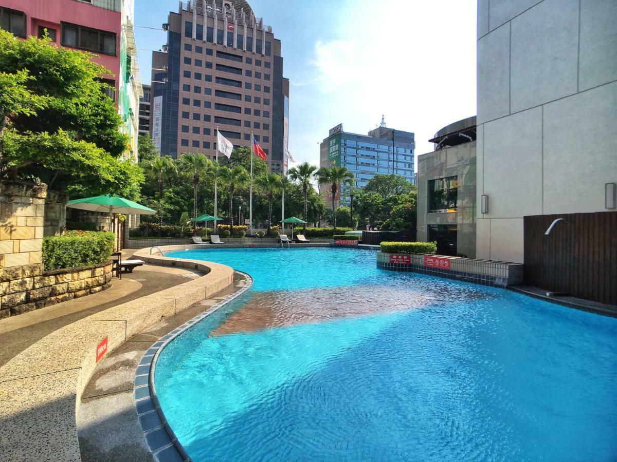 Evergreen Laurel Hotel - Taizhong Zewnętrze zdjęcie The photo shows a swimming pool set in a modern urban environment. The pool has a curved edge and is surrounded by greenery, including trees and umbrellas for shade. In the background, there are several tall buildings, indicating a cityscape, with so