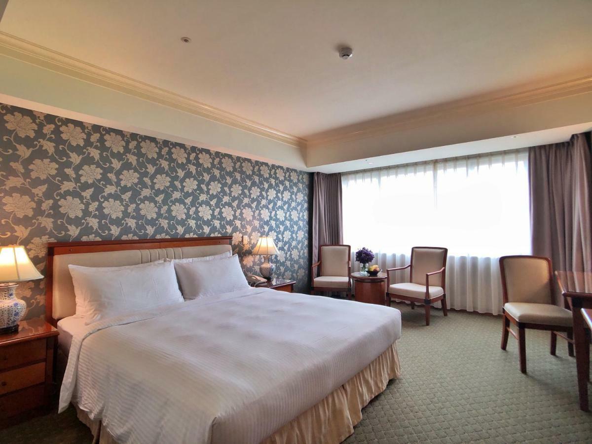Evergreen Laurel Hotel - Taizhong Zewnętrze zdjęcie The photo shows a hotel room that features a large bed with a white bedspread positioned against a decorative floral wallpaper. There are two bedside lamps on wooden nightstands flanking the bed. To the side of the room, there are two chairs and a sm