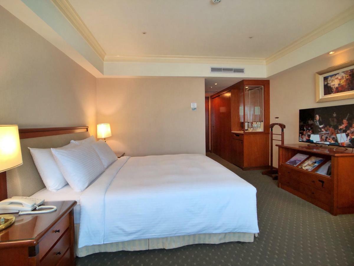 Evergreen Laurel Hotel - Taizhong Zewnętrze zdjęcie The photo shows a hotel room featuring a comfortable bed with white linens and pillows, positioned against a light-colored wall. There is a wooden bedside table with a lamp and an open book next to the bed. On the opposite side, there’s a large flat-