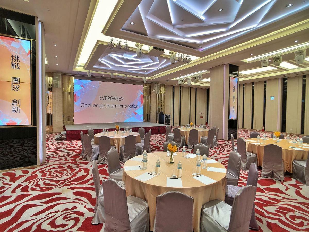 Evergreen Laurel Hotel - Taizhong Zewnętrze zdjęcie The photo shows a conference or event space set up for a gathering. The room features several round tables with light-colored tablecloths and chairs, each with decoration, possibly for dining or discussion. A large screen displays the text "EVERGREEN