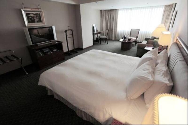Evergreen Laurel Hotel - Taizhong Zewnętrze zdjęcie The photo shows a hotel room featuring a large bed with white bedding. There is a flat-screen television on a wooden cabinet opposite the bed. Nearby, there are two armchairs and a small table, creating a cozy seating area. Large windows allow natura