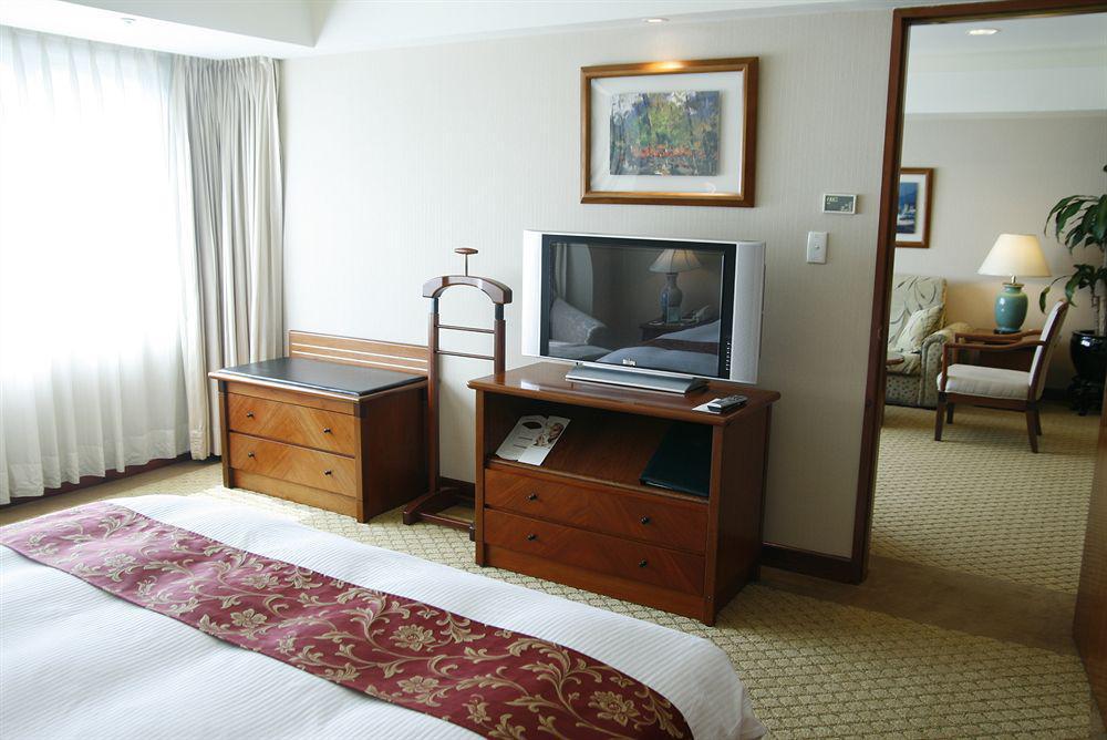 Evergreen Laurel Hotel - Taizhong Zewnętrze zdjęcie The photo depicts a hotel room interior. In the foreground, there is a neatly made bed with a decorative red bedspread. To the left, a large window allows natural light to fill the room, with light-colored curtains drawn to one side. 

Next to the be