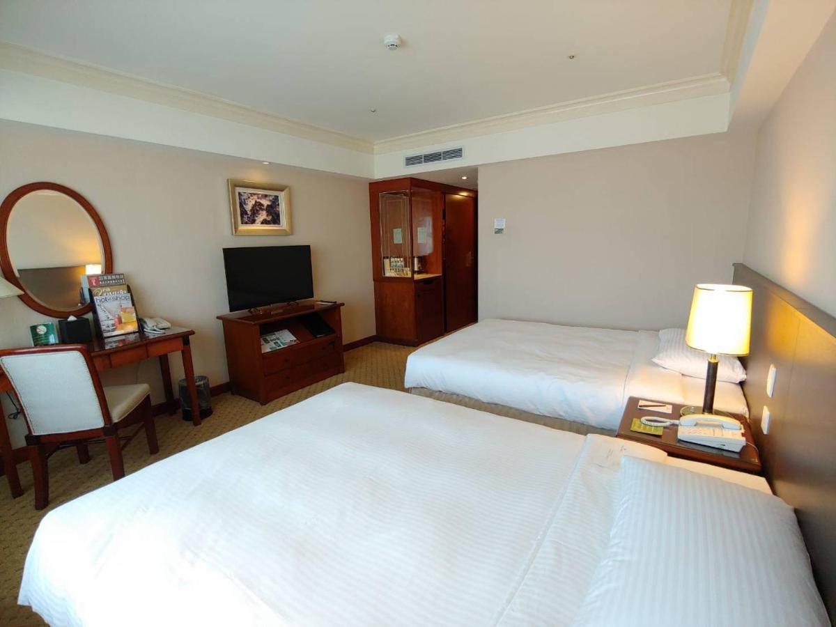 Evergreen Laurel Hotel - Taizhong Zewnętrze zdjęcie The photo shows a hotel room with two beds, neatly made with white linens. There is a desk and chair in one corner, accompanied by a flat-screen TV mounted on the wall. A large mirror and a wooden cabinet are also visible. The room is illuminated by 